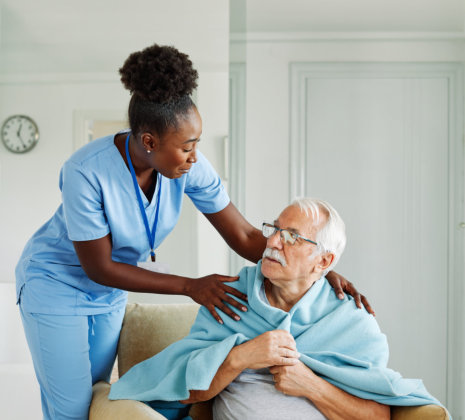 A healthcare worker in blue scrubs assists an elderly person wrapped in a light blue blanket, providing care in a home setting.