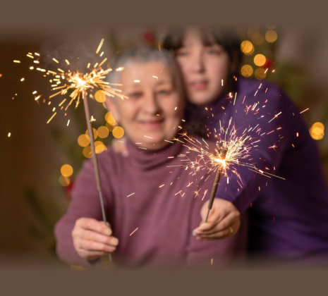 Two people holding sparklers in front of a decorated Christmas tree, celebrating in a festive indoor setting.