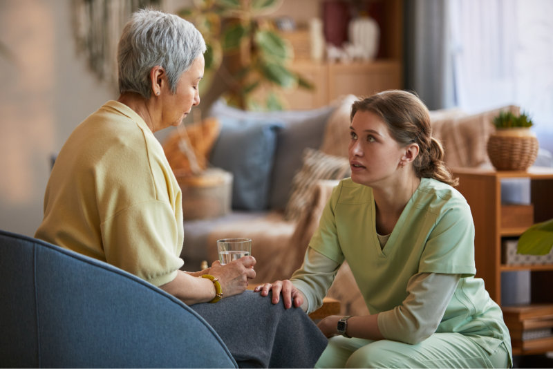 The caregiver looks up with a concerned and supportive expression, placing a hand on an elderly woman